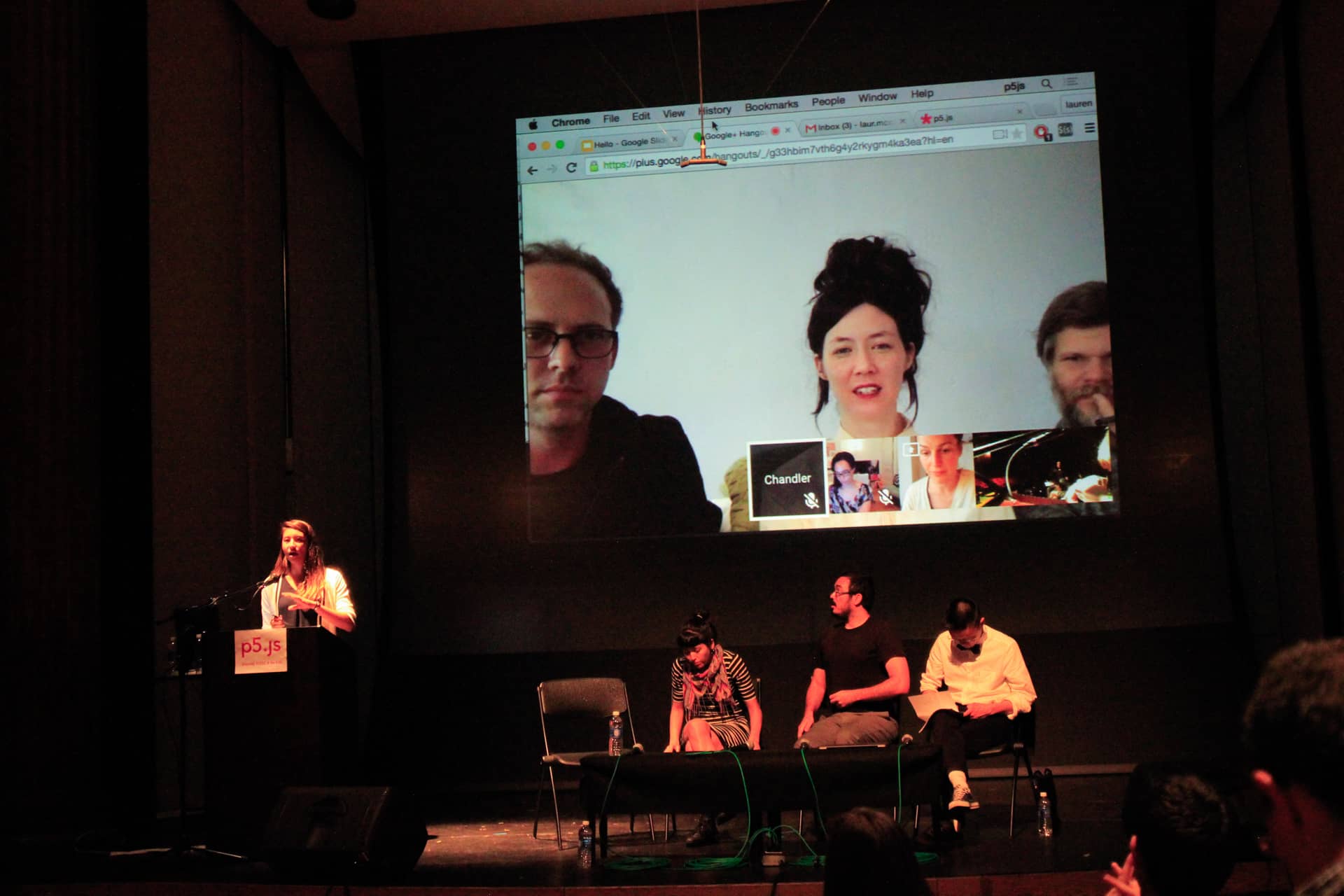 Woman speaks at a podium in an auditorium while three participants sit on the stage and another three are skyping in on the stage screen"