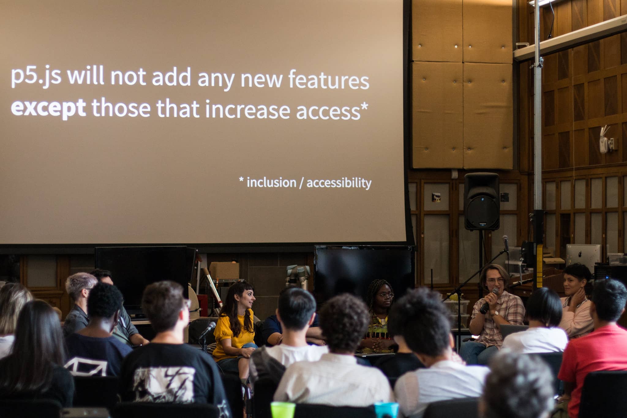 Person with a microphone speaking to fellow participants in front of text that reads p5.js will not add any new features except those that increase access