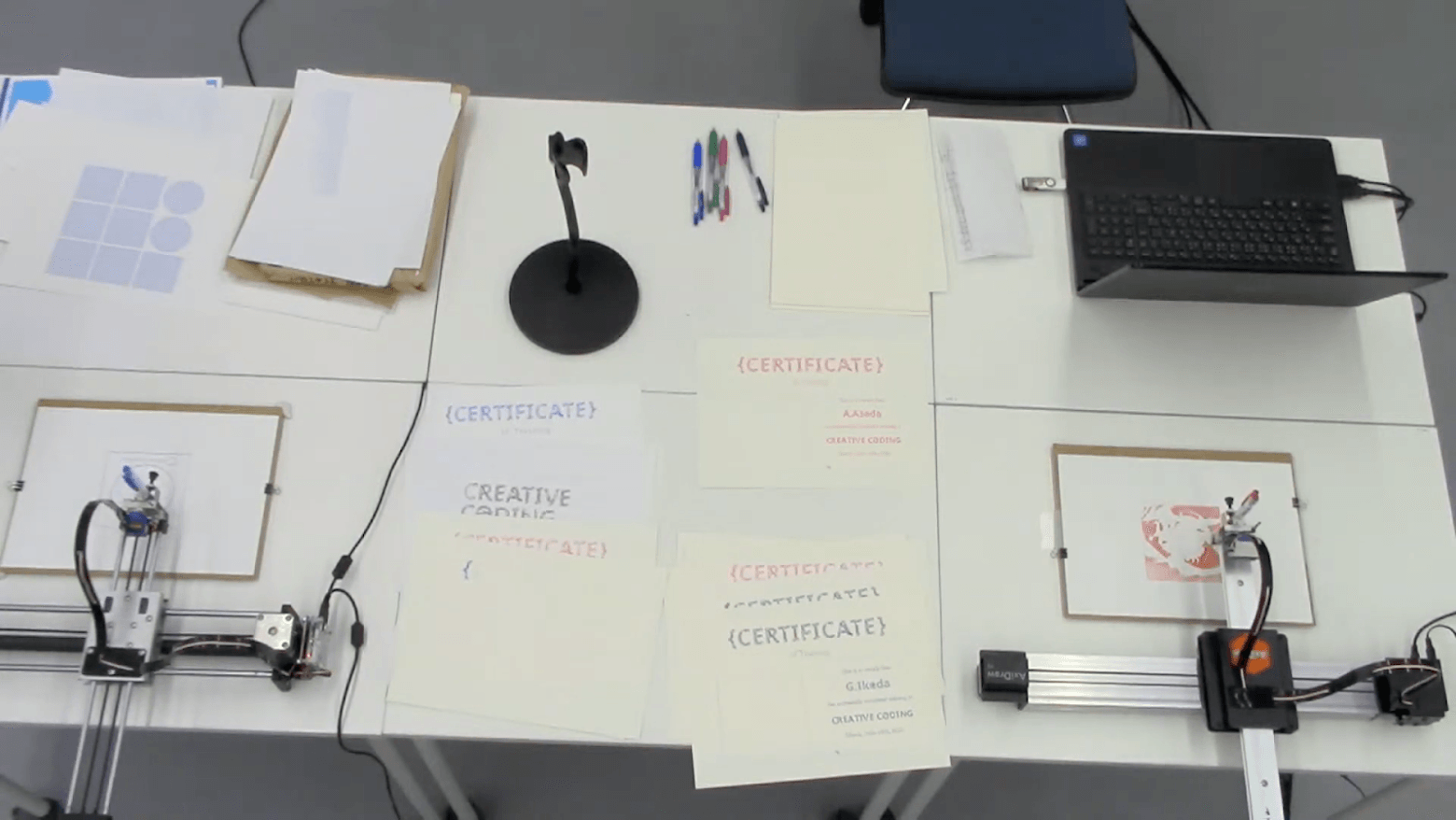 A table on which there is a laptop, some sheets of papers, colorful pens and two automatic machines drawing something with a pen on a sheet.