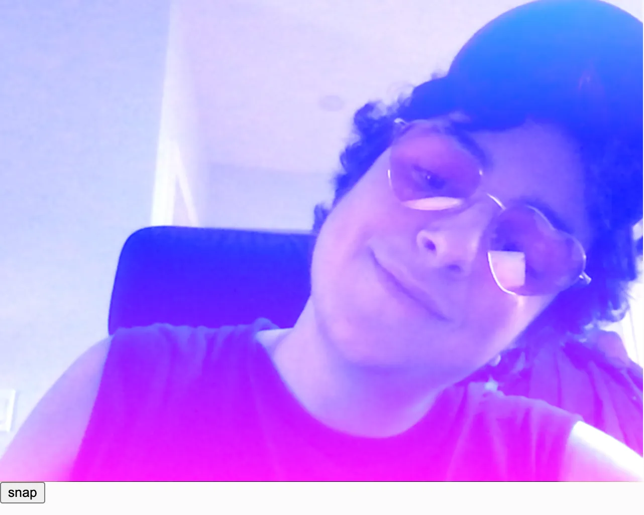 A webcam snapshot of a white person with short dark curly hair wearing pink heart shaped glasses posing for a picture. There is a red to yellow linear gradient filter on top of the photo. Colorful circles are scattered on top of the image with a color gradient overlay. The circles have randomized radial color gradients inside them to mimic a camera's lens flare. Some color gradients are pink to purple, cyan to green, or yellow to orange.