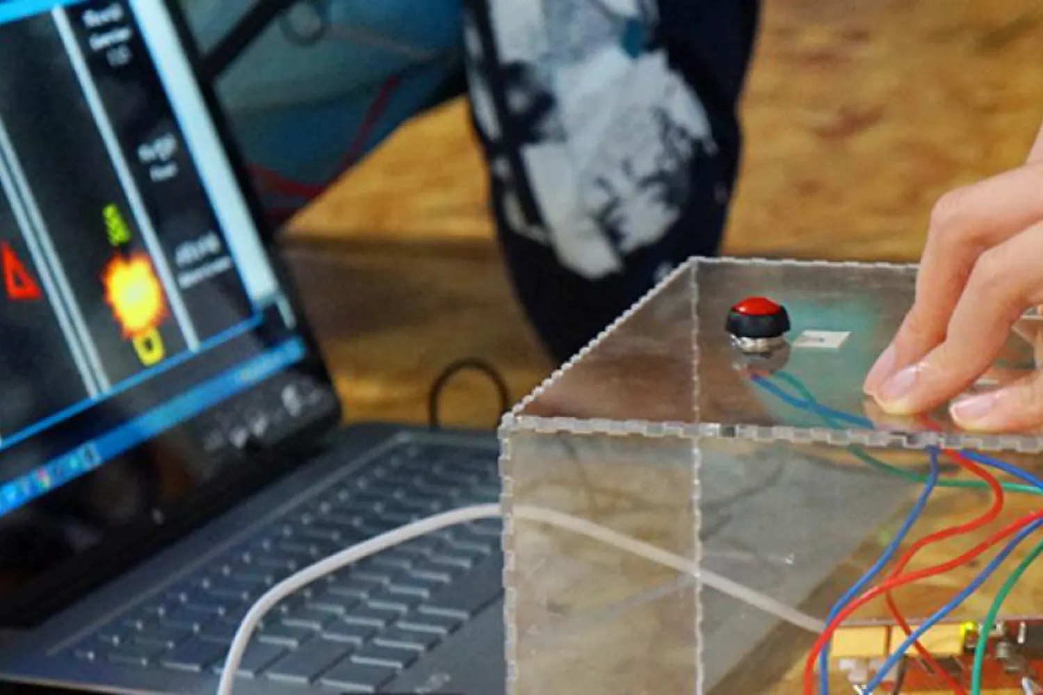 Fingers resting on an acrylic cube sporting buttons to control a game unfolding on a laptop screen visible in the background