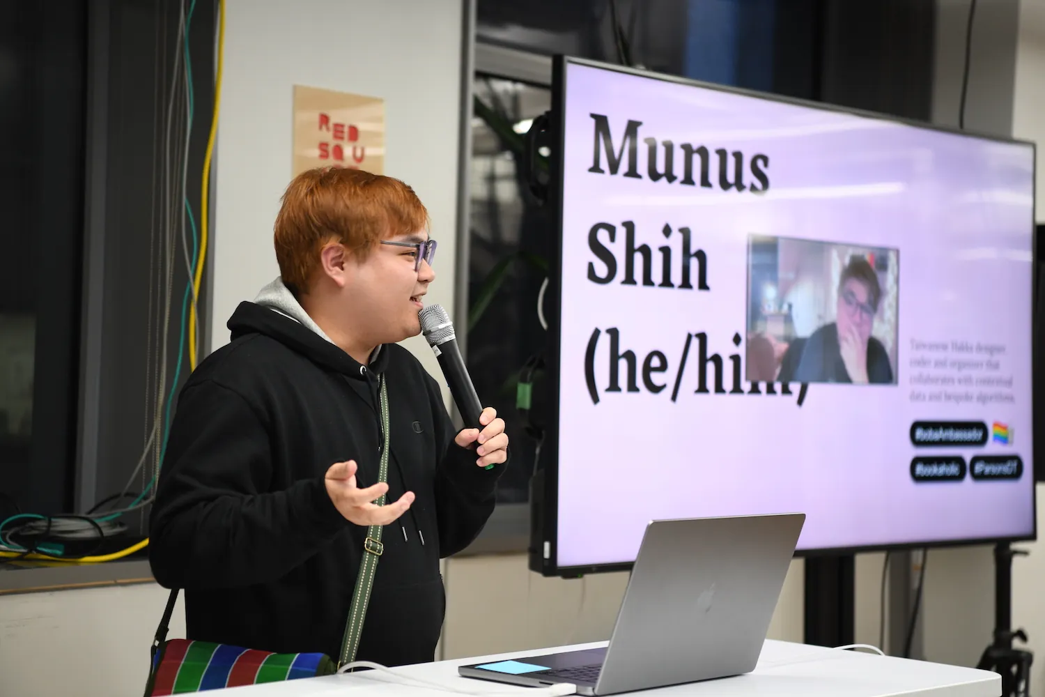 Munus Shih holding a mic next to a big t.v. screen that has an image of himself with his name, pronouns, and other facts about him.