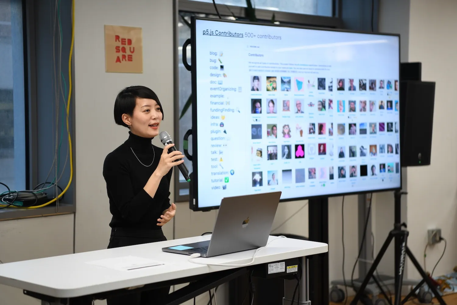 Qianqian Ye holding a mic next to a big t.v. screen that has a grid of p5.js contributor photos on it.