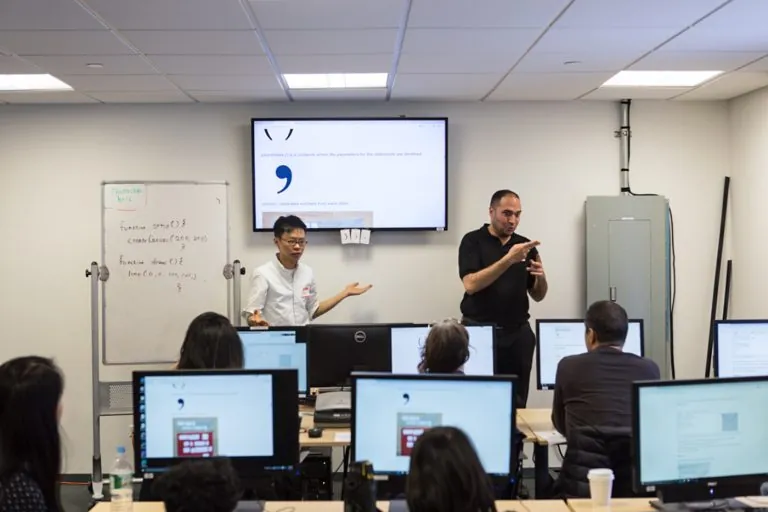 Two volunteers explaining concepts using a white board and a screen to a bunch of deaf and hard of hearing students, each student facing a computer screen.