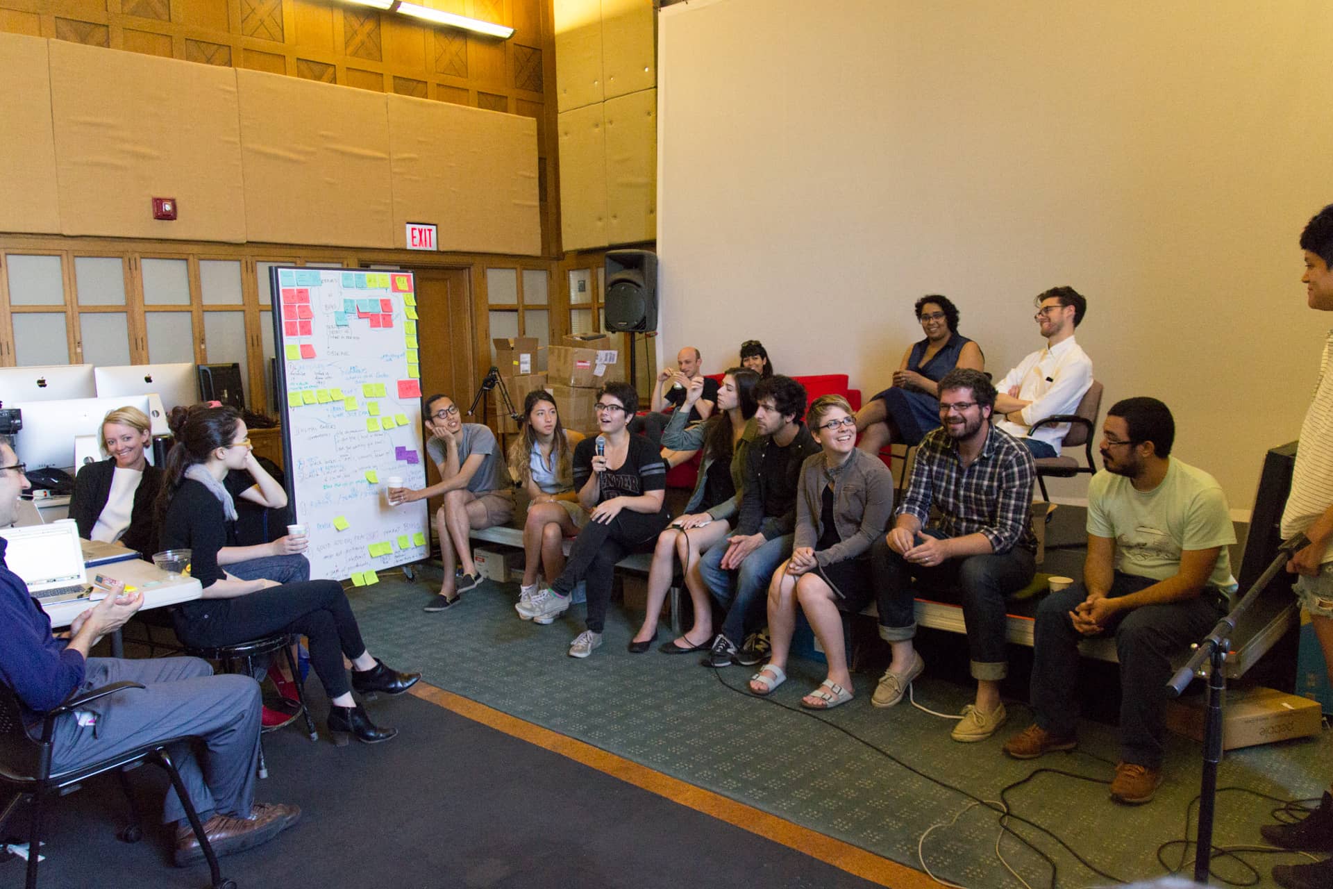 Participants sit in a circle around a white board with sticky notes on it while a female student speaks into a microphone"