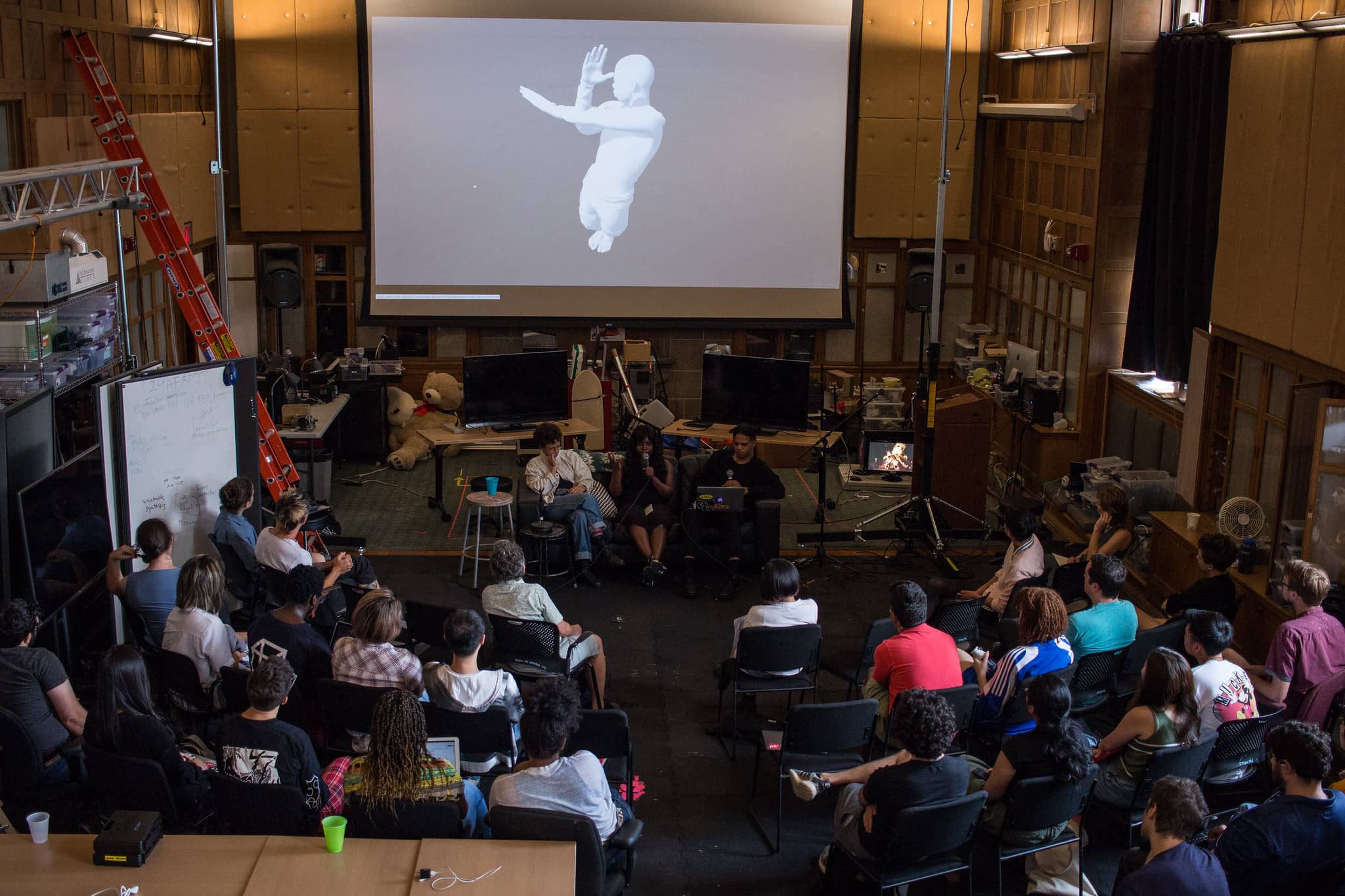 Overhead view of participants listening to a panel of people with an image of a 3d rendered man on it"